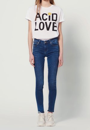 Personne portant un t-shirt blanc avec un visage souriant, un jean skinny bleu et des baskets blanches, debout devant un fond clair.