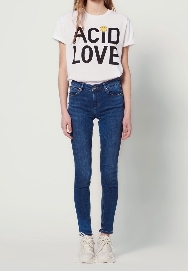 Personne portant un t-shirt blanc avec un visage souriant, un jean skinny bleu et des baskets blanches, debout devant un fond clair.