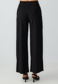 Zwarte wijde broek met dunne verticale witte strepen, gedragen met zwarte open teen hoge hakken op een witte achtergrond.