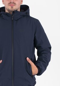 Man in een marineblauwe gewatteerde hooded jacket met rits en handen in de zijzakken tegen een effen achtergrond.