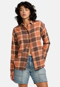 Chemise à carreaux dans des tons orange, marron et beige, en tissu texturé, avec un devant à boutons et une coupe décontractée. Associée à un short en denim.