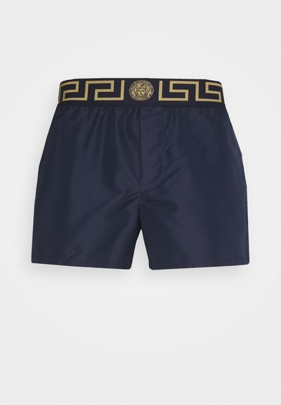 Námorné plavky s modrou farbou a zlatým gréckeho kľúča vzorom na pase, obsahujúce medailón s logom a ľahký, hladký materiál.