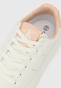 Hvit sneaker med beige tunge og krage, hvite lisser, "cK"-logo på tungen, og størrelse 33 markert på innersålen.