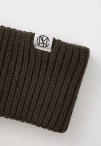 Dark green ribbed knit beanie with a small white label featuring a circular logo. The texture is soft and elastic.
