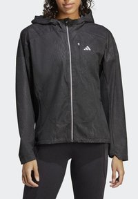 Giacca impermeabile nera con tessuto testurizzato, zip completa, cappuccio regolabile e due tasche frontali. Presenta un logo riflettente sul petto.