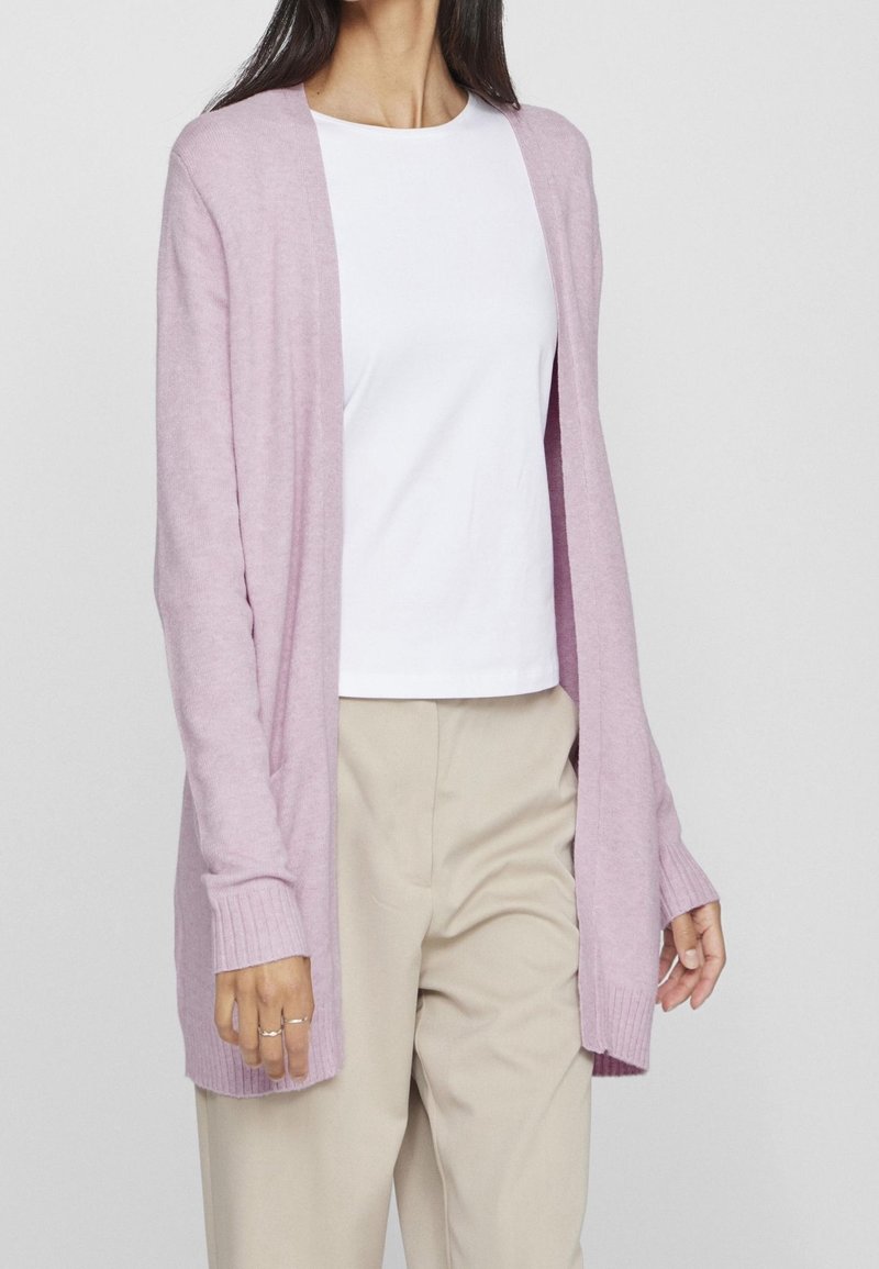 Femme portant un cardigan ouvert de couleur violet clair, un t-shirt blanc et un pantalon beige, debout devant un fond uni.
