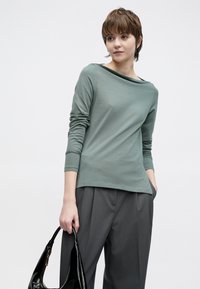 Top so dlhými rukávmi v tlmenej zelenej bavlne, s širokým výstrihom a priliehavými rukávmi, v kombinácii so sivými plisovanými nohavicami a čiernou kabelkou.