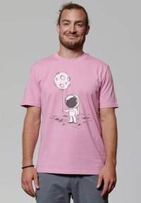 Roze katoenen t-shirt met een zwart astronautenafbeelding die een maanvormig ballonetje vasthoudt. Eenvoudige ronde hals, korte mouwen, zachte textuur.