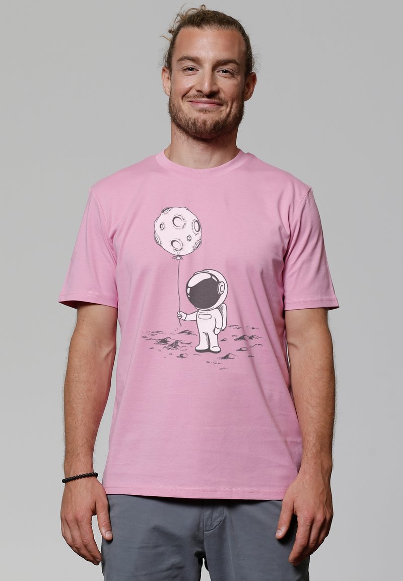 Roze katoenen t-shirt met een zwart astronautenafbeelding die een maanvormig ballonetje vasthoudt. Eenvoudige ronde hals, korte mouwen, zachte textuur.
