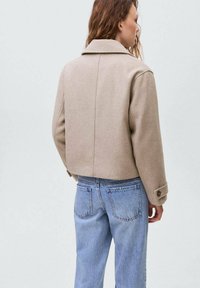 Jachetă scurtă de culoare bej cu textură subtilă, având guler ascuțit și manșete cu nasturi, purtată împreună cu blugi din denim de un albastru deschis.