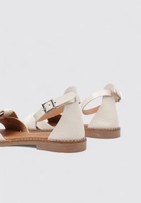 Sandales en cuir blanc cassé avec des sangles de cheville ajustables, matériau texturé, bout rond et semelle marron clair. Pas d'accents décoratifs.