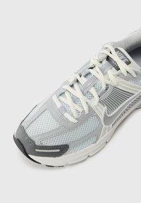 Chaussure de sport grise avec tige en mesh et superpositions synthétiques. Présente un système de laçage, un col rembourré et une semelle extérieure en caoutchouc avec une texture marquée.