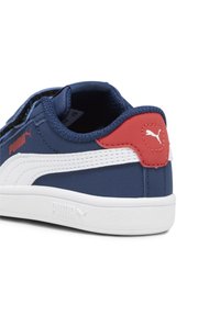 Tenis navy blue cu accente albe și toc roșu, cu un material texturat și curea reglabilă pentru o fixare sigură. Logo-ul Puma este vizibil.