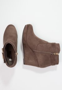 Stivaletti alla caviglia in suede marrone con tacco a zeppa, punta arrotondata, zip laterale e dettagli decorativi con cinturino. Suola in gomma piatta.