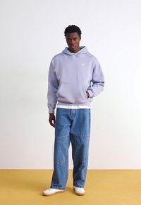 Felpa in pile viola chiaro con tasca frontale e logo bianco, abbinata a jeans in denim blu a vestibilità ampia e sneakers bianche.