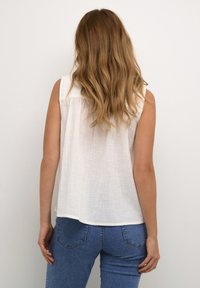 Kvinde med langt bølget hår iført en ærmeløs hvid bluse og blå jeans, der står med ansigtet mod en ensfarvet lys væg.