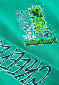 T-shirt turchese con un creeper verde pixelato di Minecraft, circondato dal testo "SSS BOOM" e "MINECRAFT" in grassetto nero.