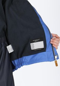 En marineblå fleecejakke med indvendigt blåt foer, der har et sort label til personlige oplysninger og plejeanvisninger samt en lynlås.