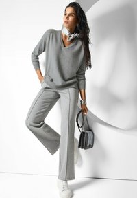 Frau in grauem V-Ausschnitt-Pullover, passenden weiten Hosen, weißen Sneakers, mit grauer Handtasche, tragt ein silbernes Armband und ein Halstuch, posiert vor einer weißen Wand.