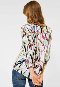 Blouse à motifs multicolores avec une coupe décontractée, des poignets élastiques et un ourlet arrière légèrement plus long, portée avec un jean en denim.