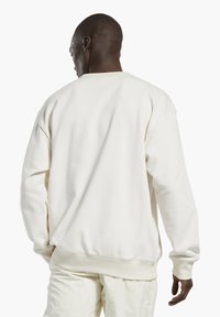 Bílá mikina se uvolněným střihem, ribbovanými manžetami a lemem a hladkým povrchem, s kulatým výstřihem a mírně spuštěnými rameny.