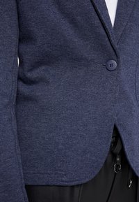 Blazer de punto azul marino con un solo botón azul, con cuello de solapa y aberturas laterales, confeccionado en una tela suave y texturizada.