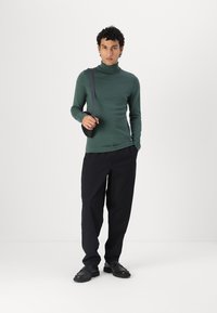 Donkergroene coltrui, slim fit, gecombineerd met losse zwarte broek en zwarte schoenen. Model houdt een zwarte schoudertas vast.