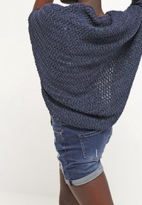 Námořnický pletený šál s volným tkaním, který se překládá přes ramena, v kombinaci s sepranými denimovými šortkami s ohrnutými manžetami.