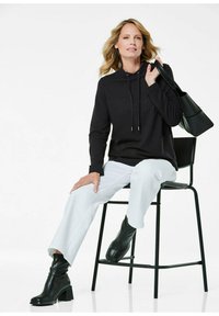 Zwarte hoodie met lange mouwen en trekkoord, gecombineerd met witte broeken. Draagt zwarte leren enkellaarsjes en houdt een zwarte shoppertas vast.