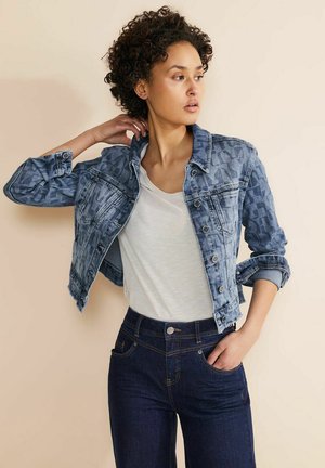 Jeansjacke aus hellblauem Denim mit einem subtilen geometrischen Muster, einreihige Knopfleiste und hochgekrempelte Ärmel, kombiniert mit einem weißen T-Shirt und dunklen Jeans.