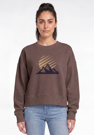 Vrouw met donker haar in een paardenstaart, die een bruin sweatshirt met berg- en zonafbeelding draagt en lichtblauwe jeans tegen een witte achtergrond.