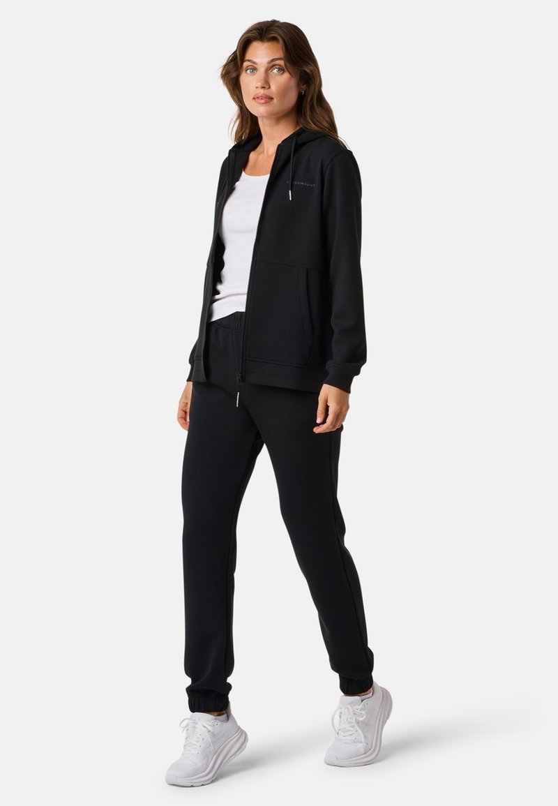 Zwarte zip-up hoodie met voorzakken, gecombineerd met bijpassende joggers; beide hebben een soepele textuur en elastische boorden. Gecombineerd met witte sneakers.