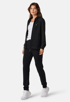 Felpa nera con zip e tasche frontali, abbinata a pantaloni jogger coordinati; entrambi presentano una texture liscia e polsini elasticizzati. Indossati con sneakers bianche.