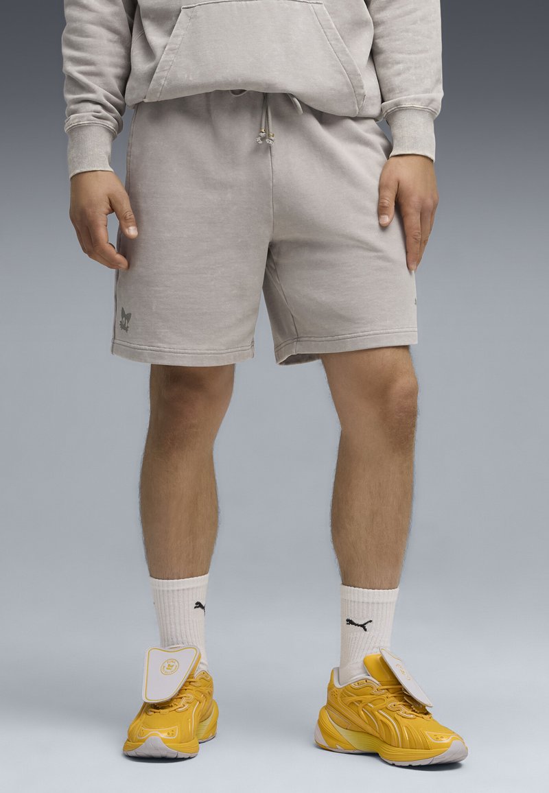 Gray cotton shorts with a drawstring waist, side pockets, and a logo detail on the left leg. Paired with yellow sneakers and white socks.