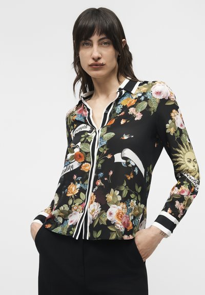 Γυναίκα που φοράει μαύρο πουκάμισο με κουμπιά, διακοσμημένο με πολύχρωμα λουλουδάτα και ηλιακά σχέδια, λευκό ριγέ γιακά και μανσέτες, με τα χέρια στις τσέπες μαύρου παντελονιού.