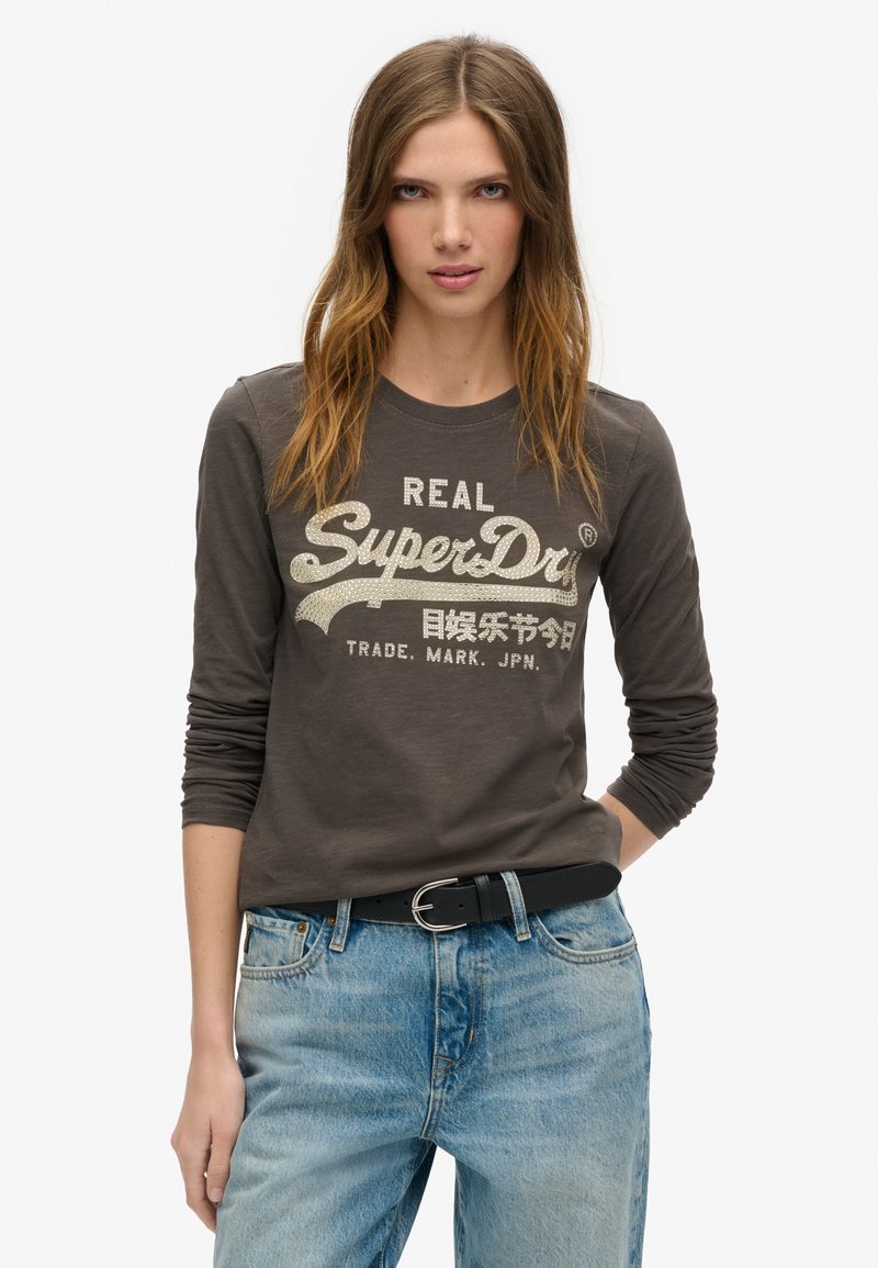 Camiseta de manga larga en gris oscuro con un logo brillante de "SuperDry". Combinada con jeans de mezclilla azul claro y un cinturón negro. Diseño sencillo y casual.