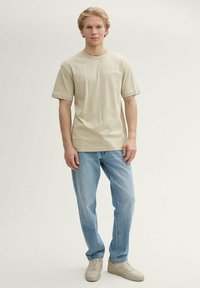 Beige katoenen t-shirt met korte mouwen, ronde halslijn en kleine grafische tekst. Gecombineerd met lichtblauwe jeans en beige sneakers.