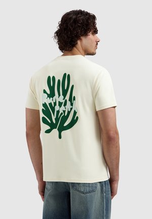Man met krullend haar draagt een crèmekleurig T-shirt met een groen abstract plantenontwerp en witte tekst op de achterkant, gecombineerd met blauwe spijkerbroek.