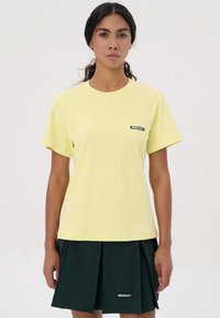 Geel katoen t-shirt met een ronde hals en korte mouwen, voorzien van een zwart logo-patch op de linkerkant van de borst. Gecombineerd met een donkergroene geplooide rok.