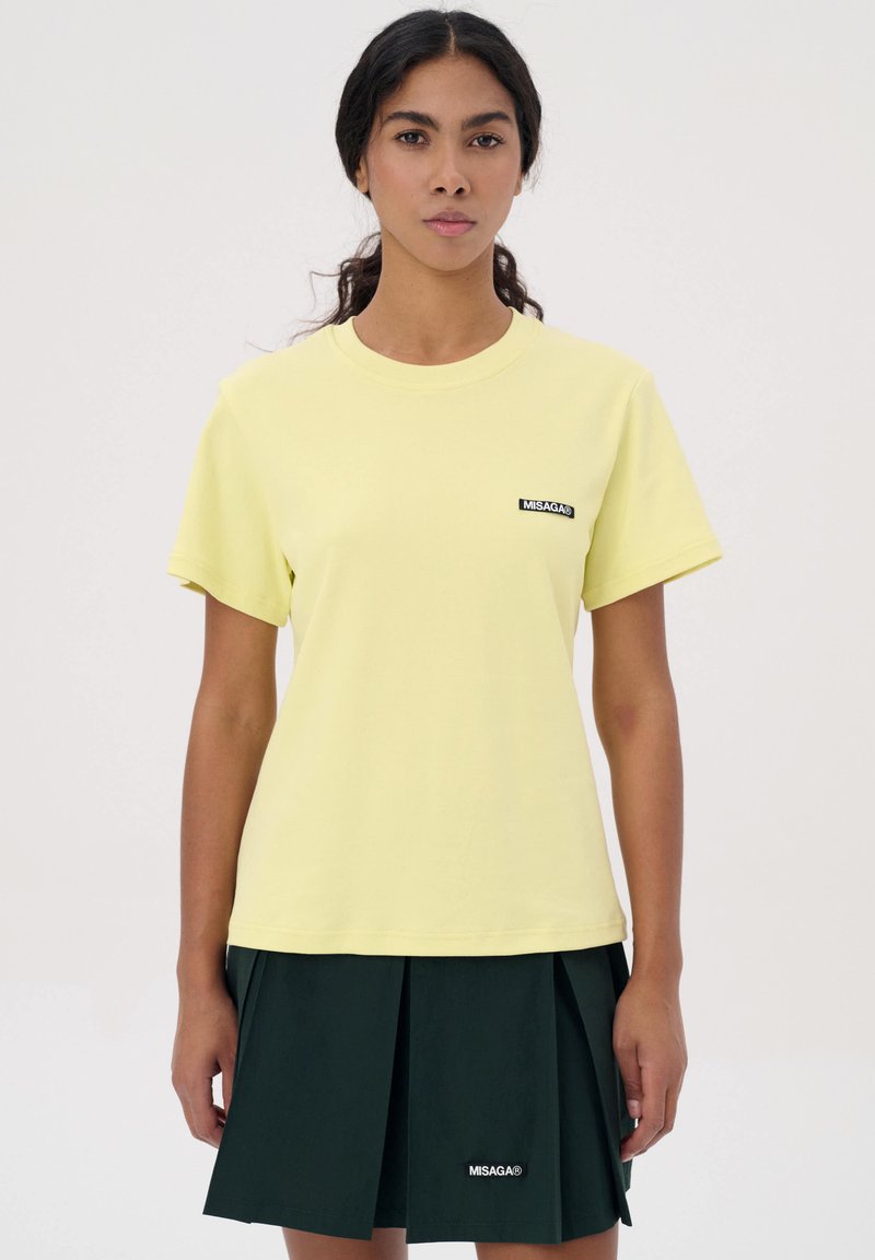 Geel katoen t-shirt met een ronde hals en korte mouwen, voorzien van een zwart logo-patch op de linkerkant van de borst. Gecombineerd met een donkergroene geplooide rok.