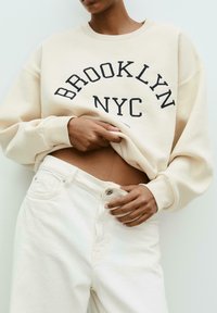 Person with medium skin tone lifting cream sweatshirt that says "Brooklyn NYC," wearing high-waisted off-white jeans unbuttoned at waist.