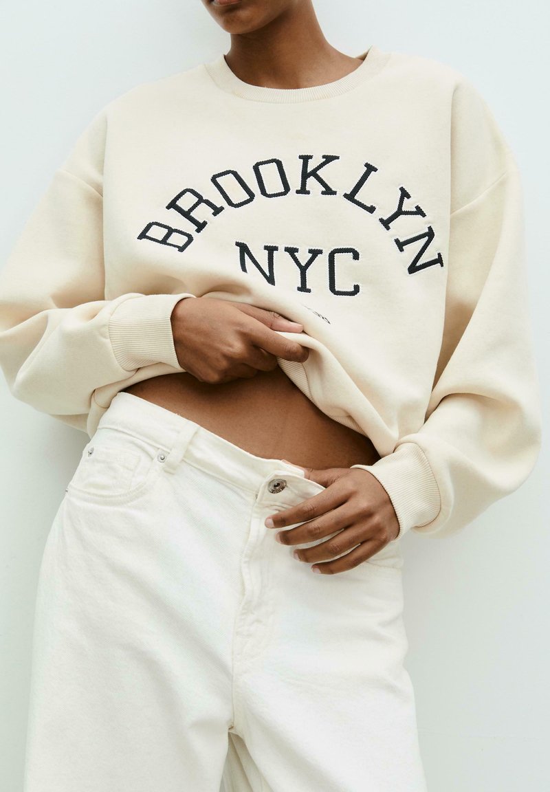 Person with medium skin tone lifting cream sweatshirt that says "Brooklyn NYC," wearing high-waisted off-white jeans unbuttoned at waist.