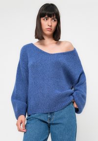 Giovane donna in un ampio maglione di lana blu con una spalla scoperta e jeans blu, in piedi contro uno sfondo chiaro uniforme.