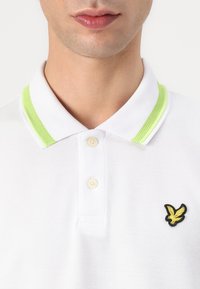 Polo bianco con tessuto a trama, caratterizzato da un colletto verde lime e un piccolo logo ricamato di un'aquila gialla sul petto.