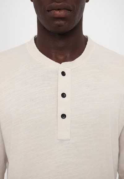 Lysebeige henley skjorte laget av teksturert stoff, med rund hals, fire svarte knapper og ribbestrikket krage.