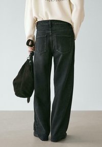 Persona che indossa jeans neri a gamba larga a vita alta e un maglione crema, con una borsa nera tenuta di fianco, in piedi contro uno sfondo neutro.