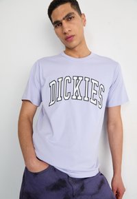 Krátký rukáv tričko levandulové barvy s nápisem "DICKIES" vytištěným tučným černě obrysem. Hladká tkanina, kulatý výstřih, nošené s šortkami s batikováním.