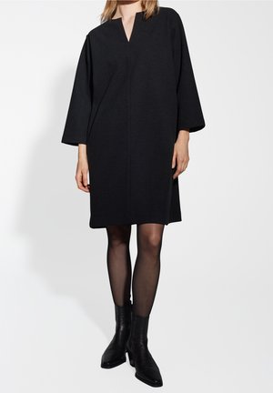 Woman wearing a knee-length black dress with wide sleeves, sheer black tights, and black ankle boots against a plain white background.