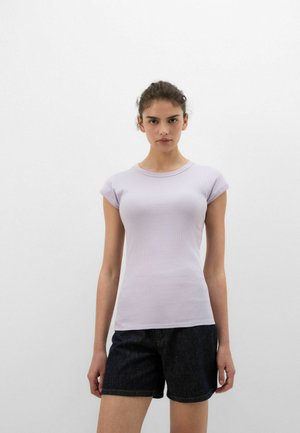 Kvinde stående mod en hvid baggrund iført en lilla kortærmet tætsiddende top og mørke denimshorts, ser frem med et neutralt udtryk.