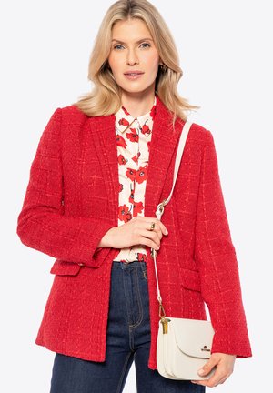 Rode blazer met textuur en twee zakken aan de voorkant, gecombineerd met een blouse met bloemenpatroon en donkerblauwe jeans, met een crèmekleurige crossbodytas.